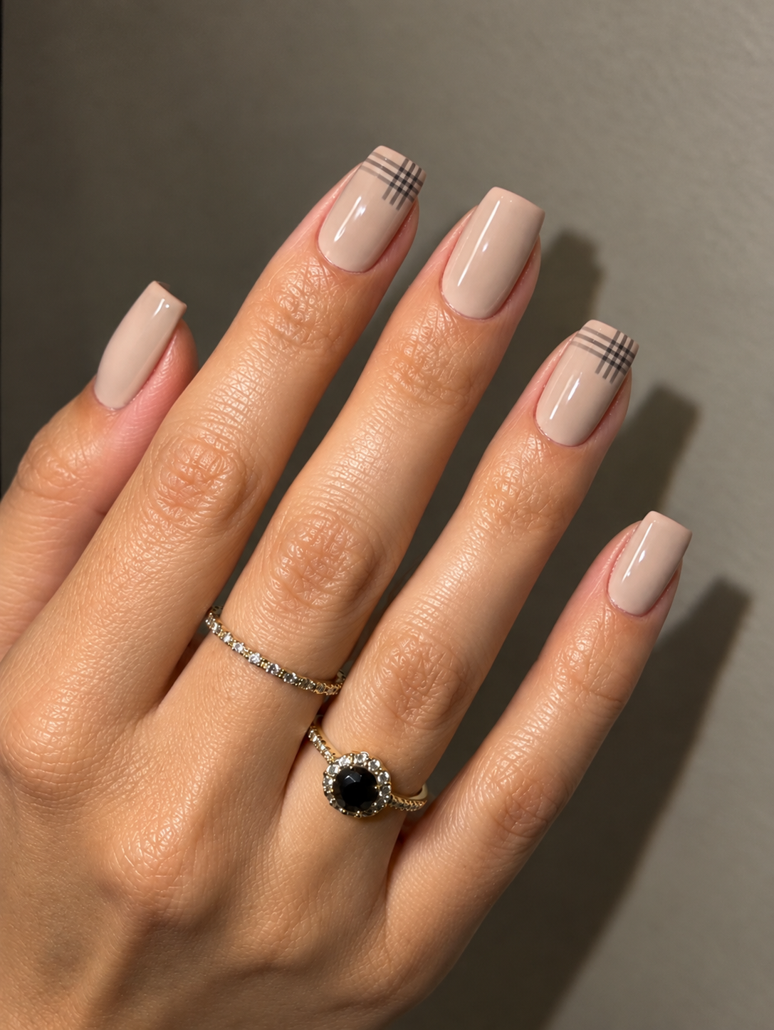 Sand-beige nails with small charcoal checked corners on the index and ring fingers.