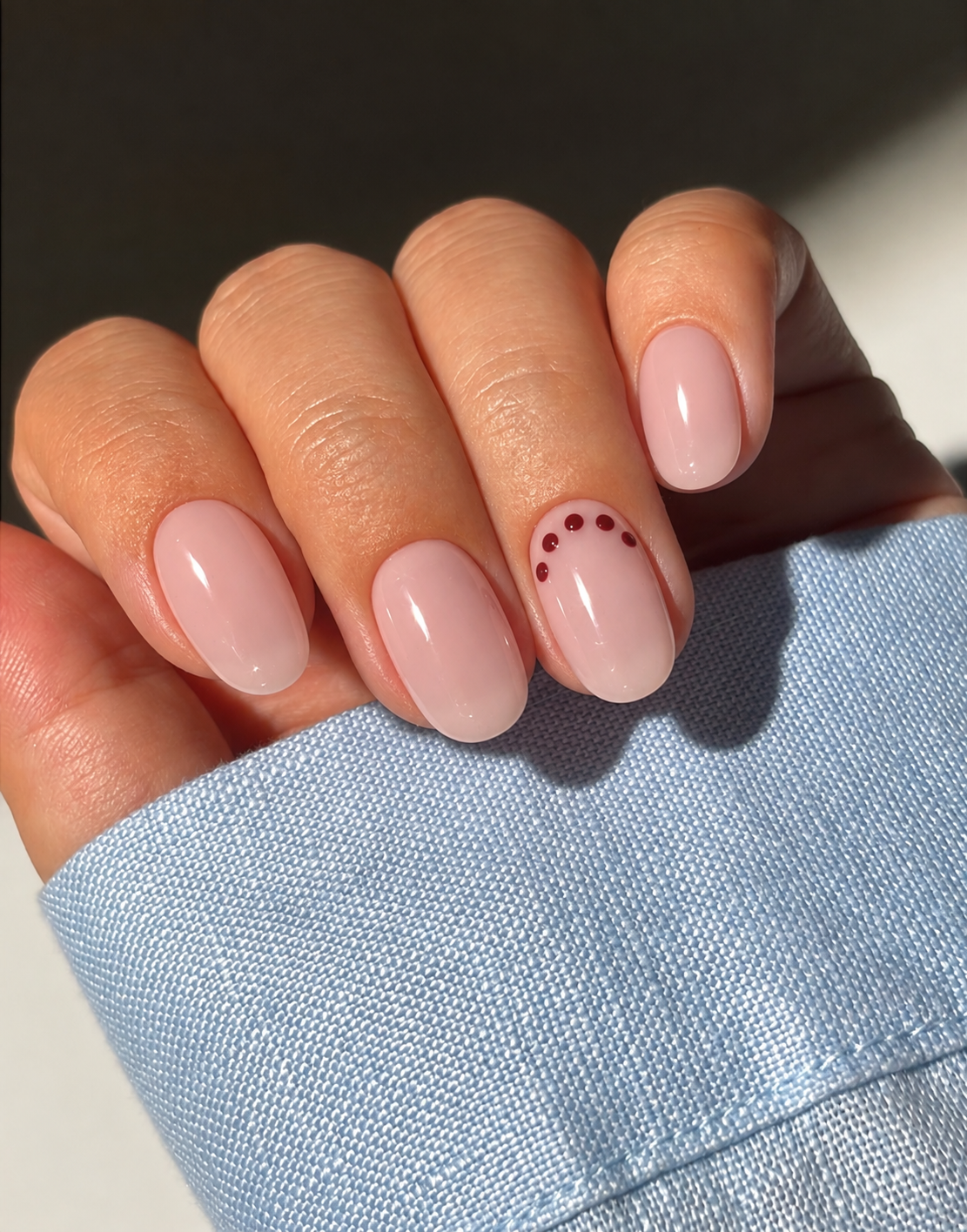 Short glossy blush nails with an arc of five cranberry dots near the ring-finger base.